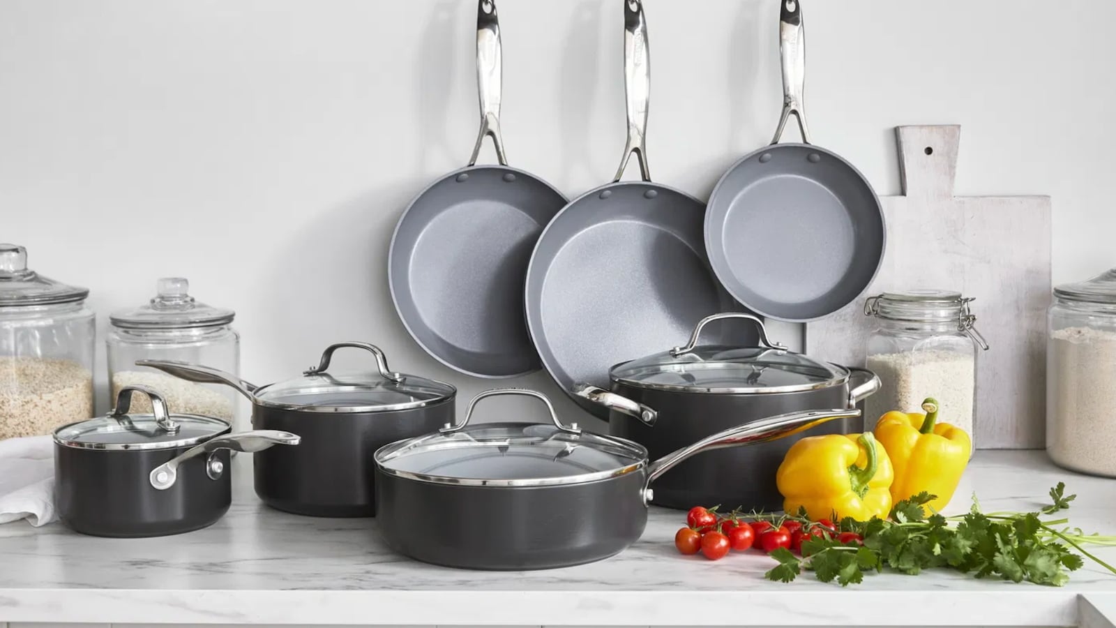 A collection of GreenPan's eco friendly cookware on a kitchen counter. There are three frying pans hanging on the wall and four saucepans on the counter. Next the pans are yellow peppers, cherry tomatoes, and parsley.